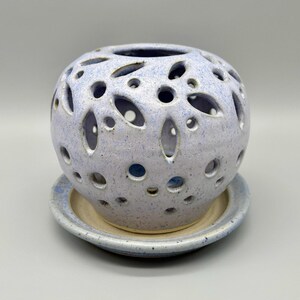 May include: A ceramic candle holder with a light blue speckled finish. The spherical holder has numerous oval and round cutouts, sitting on a matching saucer. The design allows light to filter through, creating a decorative effect.