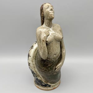 May include: A ceramic mermaid sculpture with a textured, earthy finish. The figure has long hair and is posed looking upward, with one hand on her chest. The lower half transitions into a tail with dark and light patterns.