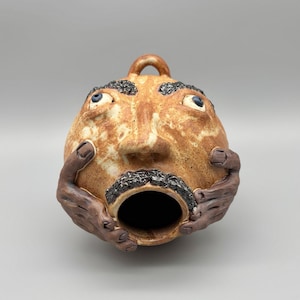 May include: A round ceramic vessel featuring a sculpted human face with a tan speckled glaze. The figure has dark textured eyebrows and a mustache, blue eyes, and two dark brown sculpted hands cupping the sides of the open circular mouth.