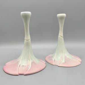 May include: Two ceramic candle holders. Each has a tall, slender, off-white stem that flares into a petal-like base. The base transitions from off-white to a soft pink. The candle holders are designed to resemble flowers.