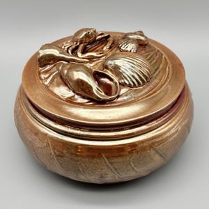 May include: A round ceramic lidded jar with a metallic copper glaze. The lid features a relief sculpture of a crab and various seashells, including a conch and a scallop shell, all finished in the same reflective, warm-toned copper finish.