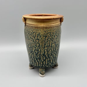 May include: A handcrafted ceramic planter with a textured, mottled surface in shades of green and brown. The rim is a lighter tan color, and the planter has three small feet. The planter has two small handles on the rim.