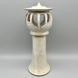 May include: A cream-colored ceramic lantern with a tall, cylindrical base and a decorative top. The lantern has cut-out designs on the globe-shaped upper section. The overall height is approximately 18 inches.