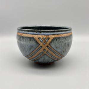 May include: A handmade ceramic bowl with a blue-grey glaze. The bowl features a band of tan-colored geometric designs around the middle. The bowl is round with a slightly flared rim and a dark brown base.