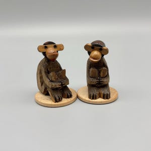 May include: Two carved wooden monkey figurines, each featuring a dark brown finish and a lighter brown face. The monkeys are depicted in a seated position with hands held near their chests, and each is mounted on a circular light wood base.