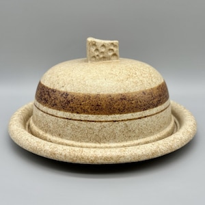 May include: A speckled, beige ceramic butter dish with a domed lid and a circular base. The lid features a textured handle and a wide, brown band. The dish is designed to hold and serve butter.