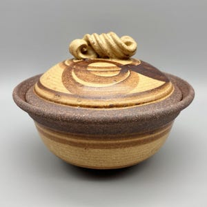 May include: A handcrafted ceramic bowl with a lid. The bowl is a light tan color with brown stripes and a dark brown rim. The lid has a swirl design and a decorative handle. This pottery piece is ideal for serving or display.