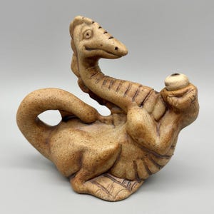 May include: A ceramic sculpture of a whimsical, light brown dragon. The dragon is in a reclining pose, with its head and tail raised. The sculpture has a textured surface and detailed features, including scales and a small, round object held in its hand.