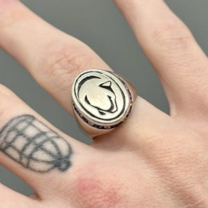 May include: Silver-toned oval ring featuring a stylized image of a cougar's head. The ring has a band of small blue stones around the oval. The ring is worn on a finger.