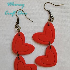 May include: A pair of red heart-shaped dangle earrings with a layered design. The earrings are made of polymer clay and have a bronze-colored hook.