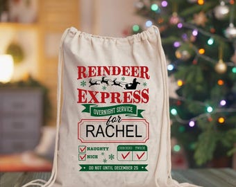May include: A cream-colored drawstring gift bag with the text "REINDEER EXPRESS" in red and green. The bag is personalized "for RACHEL" and includes a checklist for "NAUGHTY" and "NICE". The bag is decorated with snowflakes and a reindeer sleigh design.