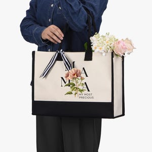 May include: A canvas tote bag with black handles and a black bottom panel. The bag features the word "MAMA" in black letters, a floral illustration, and the text "MY MOST PRECIOUS". The bag is filled with flowers.