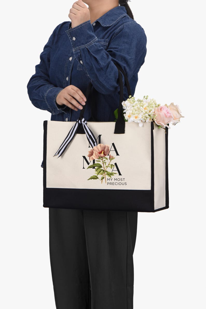 May include: A canvas tote bag with black handles and a black bottom panel. The bag features the word "MAMA" in black letters, a floral illustration, and the text "MY MOST PRECIOUS". The bag is filled with flowers.