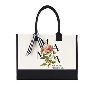 May include: A tote bag with a white background and black trim. The bag features the word "MAMA" in black letters, with a floral illustration and the words "MY MOST PRECIOUS". The handles are black with a striped ribbon.
