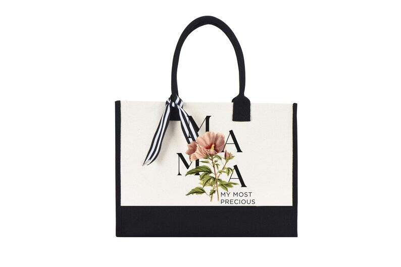 May include: A tote bag with a white background and black trim. The bag features the word "MAMA" in black letters, with a floral illustration and the words "MY MOST PRECIOUS". The handles are black with a striped ribbon.