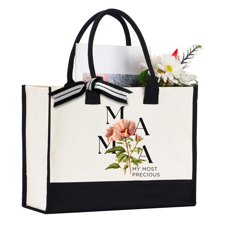 May include: A white and black tote bag with black handles and a black and white striped bow. The bag features the word "MAMA" with a pink flower illustration and the text "MY MOST PRECIOUS". The bag is filled with white paper, thread, and flowers.