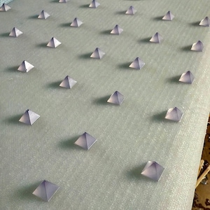 Puede incluir: Una tabla de surf blanca con una superficie texturizada cubierta de pequeños bultos piramidales de color morado. La tabla de surf tiene una pegatina ovalada marrón con la palabra "NOVA" impresa en ella.