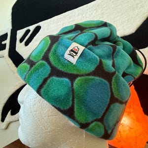 May include: A fleece beanie in shades of teal, blue, and green, with a dark brown pattern. The hat is on a white mannequin head. A small white tag with a logo is sewn on the side.