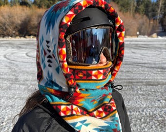 May include: A person wearing a black helmet with mirrored goggles and a patterned fleece hood. The hood features a white, turquoise, red, and yellow geometric design. The hood covers the neck and head, offering protection from the elements.