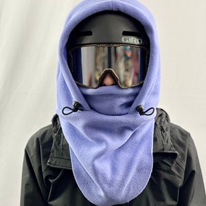 Puede incluir: Un pasamontañas de cuello de forro polar morado con cierre de cordón negro. El pasamontañas se lleva sobre un casco negro con gafas de esquí negras y plateadas.