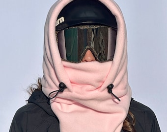 May include: A pink fleece neck gaiter with black drawstrings. The gaiter is worn over a person's head and covers their face, leaving only their eyes visible. The person is wearing ski goggles and a black helmet.