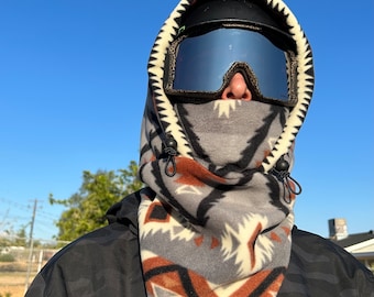 May include: A gray and brown fleece neck gaiter with a geometric pattern. The gaiter has a black drawstring and is worn over a black helmet and ski goggles.