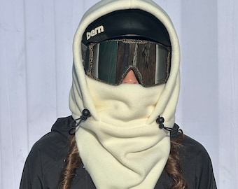 May include: Cream-colored fleece balaclava with a drawstring closure. The balaclava covers the head and neck, with a black helmet and goggles visible. The goggles have a reflective lens. The word "bern" is printed on the helmet.