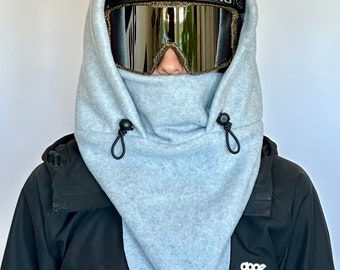 May include: A grey fleece ski mask with a drawstring closure. The mask is worn over a black ski helmet and goggles.