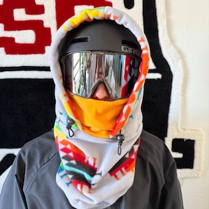 May include: A white fleece neck gaiter with a colourful geometric pattern. The gaiter has a black drawstring and is worn over a black helmet with a Giro logo. The person is wearing mirrored ski goggles.