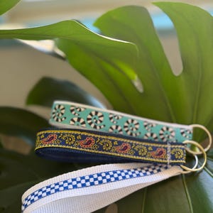 Pode incluir: Três chaveiros com padrões diferentes. O primeiro chaveiro é branco com um padrão de redemoinho preto e turquesa. O segundo chaveiro é azul marinho com um padrão paisley dourado e vermelho. O terceiro chaveiro é branco com um padrão xadrez azul e branco.