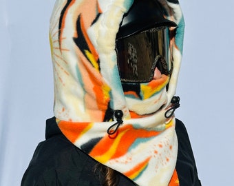 May include: A fleece balaclava with a colorful abstract pattern in orange, yellow, and teal. The balaclava has a drawstring closure and is worn with black ski goggles and a black jacket. The hood is lined with a soft, white material.