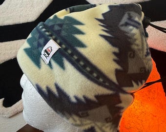 May include: A fleece beanie hat with a cream, teal, and gray geometric pattern. The hat has a small tag with a logo. The hat is on a white mannequin head. A black drawstring is visible.