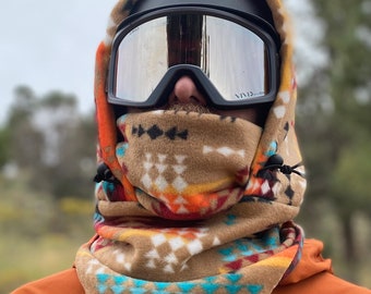Könnte beinhalten: Eine Person trägt einen braunen und blauen gemusterten Fleece-Halswärmer mit einer schwarzen Skimaske und einer Skibrille. Die Person trägt eine leuchtend orangefarbene Jacke mit dem Wort "patagonia" auf der Vorderseite.