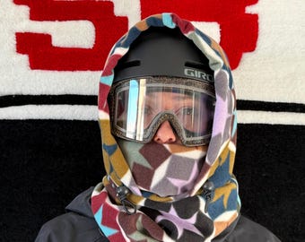 May include: A person wearing a black ski helmet with goggles and a patterned fleece hood. The hood features a geometric design in shades of brown, blue, purple, and yellow. The background is a white rug with a large red "SB" logo.