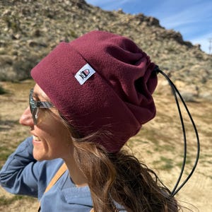 May include: A burgundy fleece beanie with a drawstring closure. The hat features a small white tag with a graphic. The beanie is worn outdoors, with a mountain backdrop.