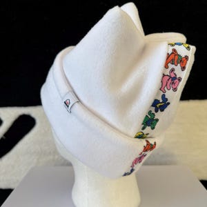 Può includere: Cappello in pile bianco con un nastro colorato con orsi danzanti. Il cappello è esposto su una testa di manichino. Una piccola etichetta è visibile sul lato del cappello. Lo sfondo è bianco e nero.