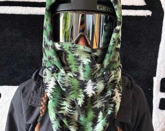 May include: A black Giro ski helmet with mirrored goggles and a green and white fleece balaclava with a pine tree design. The balaclava has black toggles and is worn over a black jacket. The image is taken indoors.