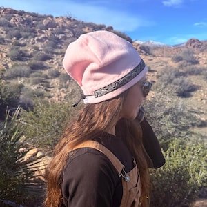 May include: A light pink beanie with a decorative black band. The hat is worn with a black long-sleeve shirt and brown overalls. The background features a desert landscape with mountains and a blue sky.
