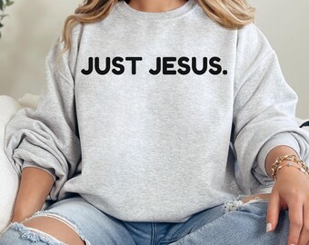 May include: A light grey sweatshirt featuring the black text "JUST JESUS.". The sweatshirt is worn with blue jeans. The image is a medium shot, focusing on the upper body.