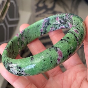 May include: A green and pink zoisite bangle bracelet. The polished stone features a vibrant green base color with speckled black and pink inclusions. The bracelet is smooth and rounded, showcasing the natural patterns of the stone.