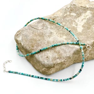 May include: A turquoise and silver necklace with a delicate design. The necklace features small, rectangular turquoise beads strung together with silver accents and a silver clasp. The necklace is displayed on a textured, light-coloured rock.