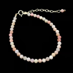 May include: A delicate bracelet featuring small, faceted beads in shades of pink, peach, and white. The beads are strung together with a silver-colored chain and clasp. The bracelet is displayed against a black background.