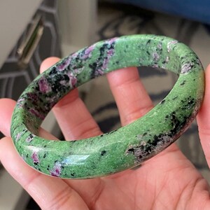 May include: A green zoisite bangle bracelet with ruby inclusions. The polished gemstone bracelet features a vibrant green base colour with black and pink speckles throughout. The bangle is held in a hand, showcasing its smooth, rounded shape.