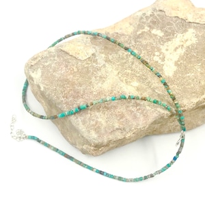 May include: A turquoise and brown beaded necklace with a silver clasp. The necklace is on a light brown rock.