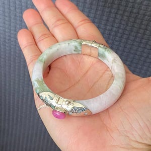 May include: A hinged bangle bracelet featuring a smooth, mottled pale green and white stone band. The bracelet includes a silver-toned metal clasp mechanism decorated with an oval-shaped pink cabochon stone and a small safety chain.