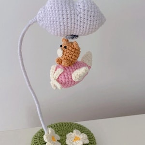 May include: A whimsical crochet lamp featuring a brown bear in a pink and white airplane suspended from a white cloud. The lamp rests on a green base with white flowers. The lamp's stem is white.