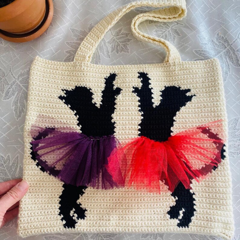 May include: A crocheted tote bag with a white background and black silhouettes of two ballerinas wearing red and purple tulle tutus.