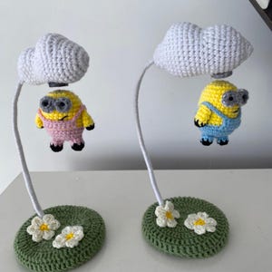 Puede incluir: Dos figuras de Minion de ganchillo, cada una suspendida de una nube blanca. Los Minions son amarillos con monos azules o rosas y gafas grises. Flotan sobre bases circulares verdes con acentos de flores blancas. El fondo es blanco.