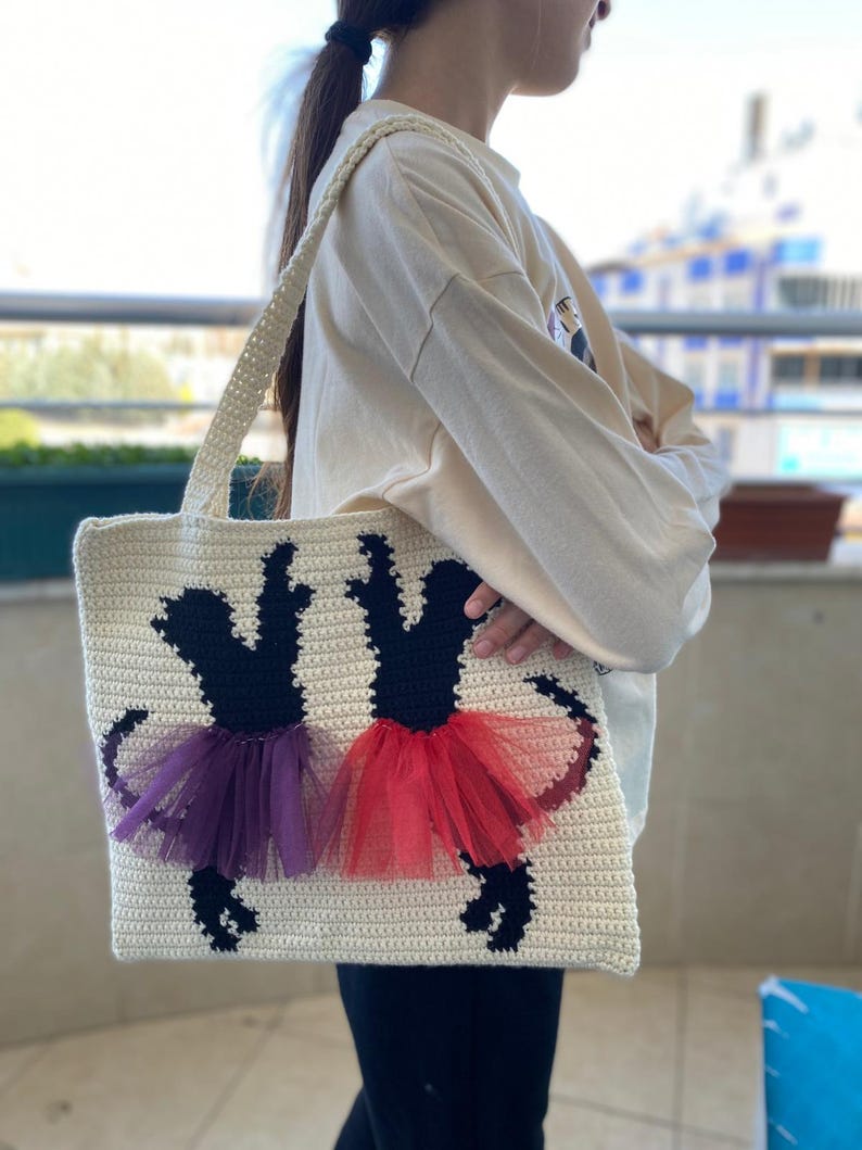 May include: A white crocheted tote bag with black silhouettes of two ballerinas wearing purple and red tutus.