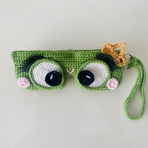 Può includere: Una maschera per gli occhi verde all'uncinetto, che assomiglia alla faccia di una rana, con grandi occhi bianchi e neri, guance rosa e una piccola corona d'oro. Ha un cinturino verde. La maschera è fatta a mano.